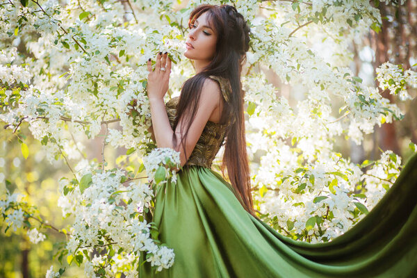 A beautiful young woman in a green dress with a large train among the blooming apple trees. Fairy princess with black long hair