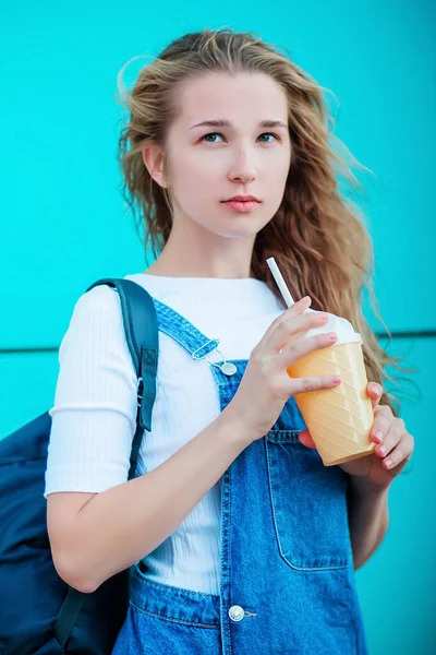 ターコイズブルーの壁を背景にした美しい少女は アイスクリームの形でコーヒーのガラスを保持しています ストリートでデニムサンドレス飲料カクテルでファッショナブルな女性 — ストック写真