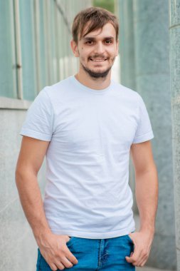 Beyaz tişörtlü genç bir adam gri bir sütunun arkasında duruyor. Şehirdeki bir ofis binasının yakınında bir adam. Model.