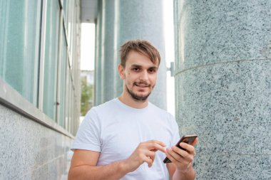 Beyaz tişörtlü genç bir adam gri bir sütunun arkasında duruyor ve akıllı telefona bakıyor. Şehirdeki bir ofis binasının yakınında elinde telefon tutan bir adam. Model.