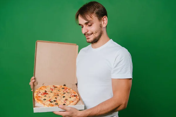 A young man in a white T-shirt holds a pizza box in his hands on a green background. Handsome guy Pizza delivery guy