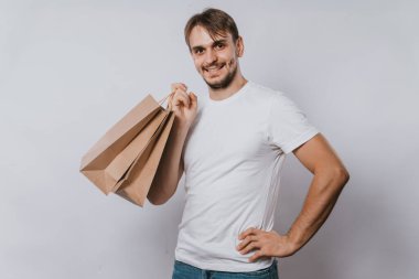 Beyaz tişörtlü genç bir adam elinde temiz kağıt torbalar tutuyor. Beyaz bir arka planda elinde alımları olan yakışıklı bir adam. Model.