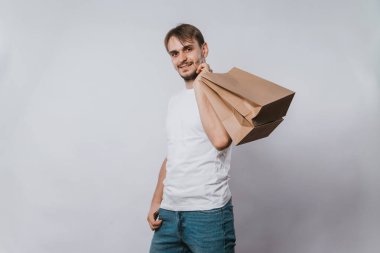 Beyaz tişörtlü genç bir adam elinde temiz kağıt torbalar tutuyor. Beyaz bir arka planda elinde alımları olan yakışıklı bir adam. Model.