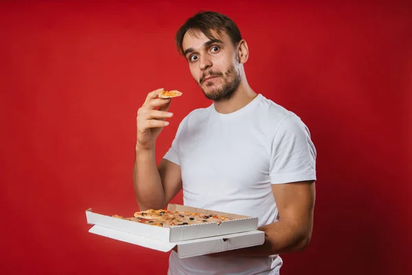 A young man in a white T-shirt holds a box in his hands and eats pizza. Handsome guy on a red background eats fast food.Mock-up.