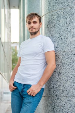 Beyaz tişörtlü genç bir adam gri bir sütunun arkasında duruyor. Şehirdeki bir ofis binasının yakınında bir adam. Model.
