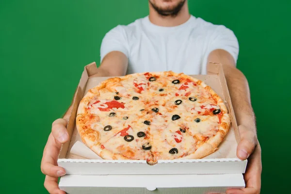 A young man in a white T-shirt holds a pizza box in his hands on a green background. Handsome guy Pizza delivery guy