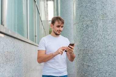 Beyaz tişörtlü genç bir adam gri bir sütunun arkasında duruyor ve akıllı telefona bakıyor. Şehirdeki bir ofis binasının yakınında elinde telefon tutan bir adam. Model.