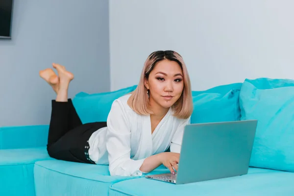 金髪のアジア系の若い女性が青いソファの上に腰を下ろしている 少女は隔離中に家で働いている 遠隔作業またはオンライントレーニング — ストック写真