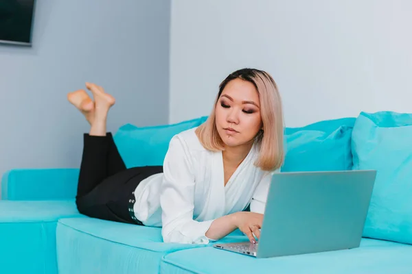 金髪のアジア系の若い女性が青いソファの上に腰を下ろしている 少女は隔離中に家で働いている 遠隔作業またはオンライントレーニング — ストック写真