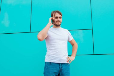 Beyaz tişörtlü genç bir adam mavi bir duvarın arkasında duruyor ve telefonla konuşuyor. Adamın elinde akıllı telefon var. Model.