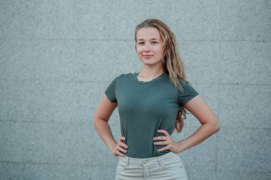 Siyah turkuaz tişörtlü genç bir kadın sokakta gri bir duvara karşı duruyor. Boş elbiseli kız. Model.