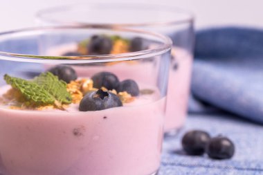 Mavi bir masa örtüsü üzerinde yaban mersini ve mısır gevreğiyle iki bardak çilekli yoğurt yakın plan.