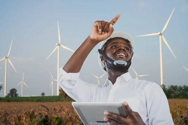 African engineer wearing white hard hat standing with digital tablet against wind turbine