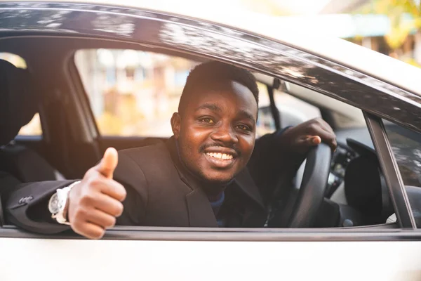 Siyah takım elbiseli Afrikalı adam gülümseyerek ve mutlu bir şekilde direksiyonda oturuyor.