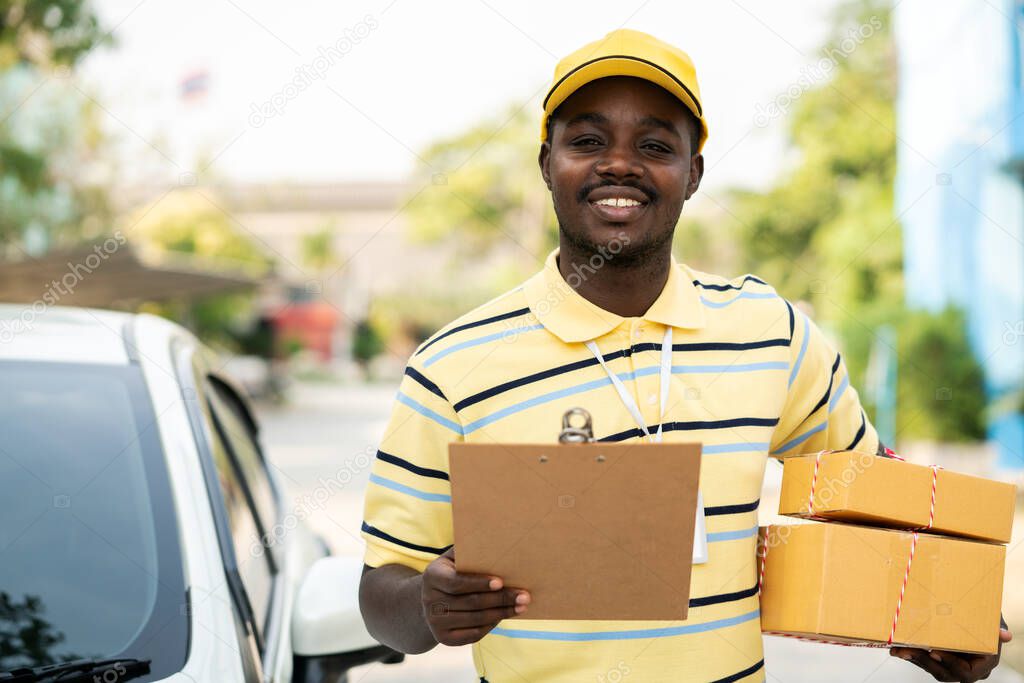 African male postal delivery courier man holding delivering package boxs and cardboard in front of car