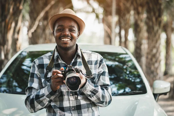 Happy traveler african man on the road with white car and holding camera