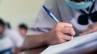 Elle kalem yazan öğrenci sınavlar yapıyor. Yüz maskesi takarak sınıfta cevap kağıtları egzersizleri yapıyor.