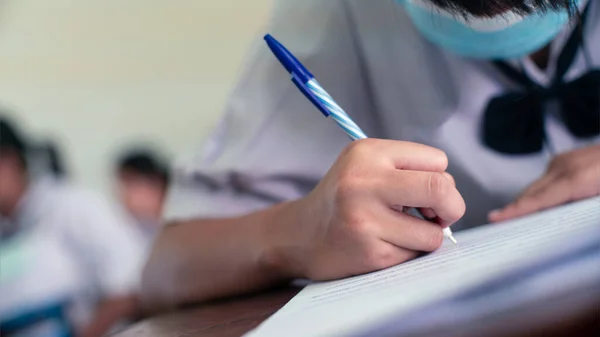 Elle kalem yazan öğrenci sınavlar yapıyor. Yüz maskesi takarak sınıfta cevap kağıtları egzersizleri yapıyor.