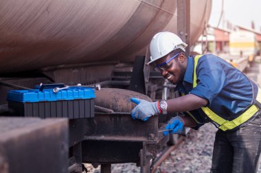 Miğfer, koruluk ve güvenlik yeleği giyen Afrikalı makine mühendisi, tren nakil gazı ve petrol nakil aracını tamir etmek için İngiliz anahtarı kullanıyor.