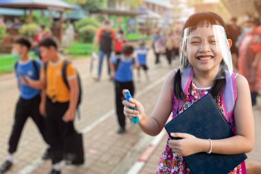 Kovid 19 karantinasından sonra okula giderken yüz maskesi takan ve elinde alkol spreyi tutan Asyalı küçük kız.