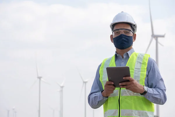 Engineer windmills  wearing face mask and hoding tablet with wind turbine in background
