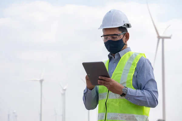 Engineer windmills  wearing face mask and hoding tablet with wind turbine in background