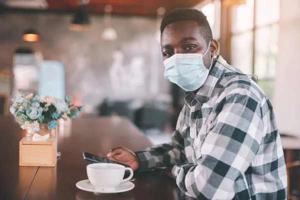 African man wearing face mask inside the coffee shop with using a smartphone