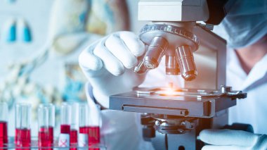 Biyoloji bilimadamı tıbbi koruyucu yüz maskesi takıyor. Mikroskop kullanarak kan örneklerine bakıyor. İlaç laboratuarında yeni aşılar için araştırma yapıyorlar.