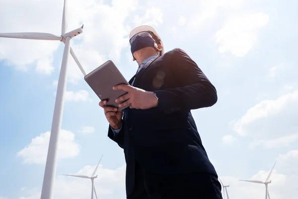 Successful engineer windmills manager wearing face mask  and using tablet with wind turbine in background