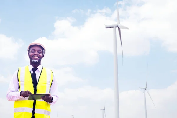 Successful african engineer windmills standing and hoding tablet with wind turbine in background