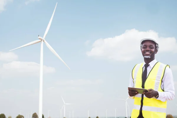 Successful african engineer windmills standing and hoding tablet with wind turbine in background