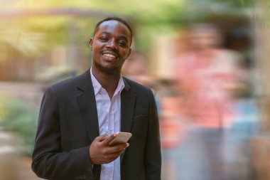 Kendine güvenen Afrikalı iş adamı akıllı telefon kullanıyor ve gülümseyerek şehirde mutlu mesut duruyor.