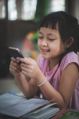 Asyalı küçük kız elinde telefon mesafesi tutarak ders öğreniyor online ders seyrediyor okullar kapalı olduğu için Covid-19
