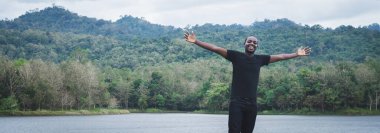 Afrikalı adam, kollarını doğa dağlarına uzatmış, gülümseme ve mutlulukla gölde duruyor.