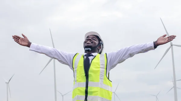 Arka planda rüzgar türbini pervanesi ve açık mavi gökyüzü olan Afrikalı erkek mühendisler çalışıyor. Alternatif enerji, gelecek için çevre dostu. temiz enerji yeniliği