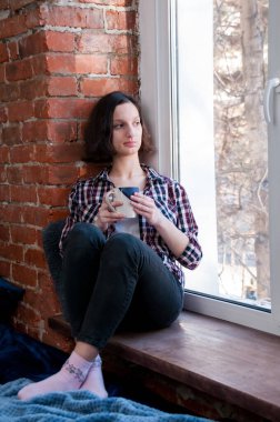 Elinde kupa olan bir kız, kışın sıcak bir evin penceresinden dikkatlice dışarı bakar.