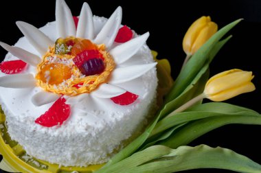 Beyaz pasta, taç yaprakları ve jöle ile süslenmiş, ve siyah arka planda sarı laleler.