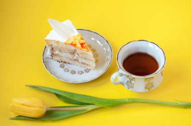 Bir fincan çay, bir tabak kek ve sarı arka planda bir lale çiçeği.