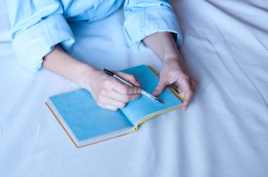 Kız, boş mavi bir defterde, yatakta duran kalemle yazıyor..