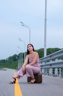 Bir kız boş bir yolun kenarında bir bavulda oturuyor ve araba bekliyor.