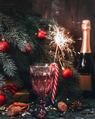 Bardakta şampanya, bardakta Noel şekeri asılı, maytap yanıyor. Arka planda Noel oyuncaklarıyla süslenmiş bir Noel ağacı ve bir şişe şampanya var. Yeni Yıl.