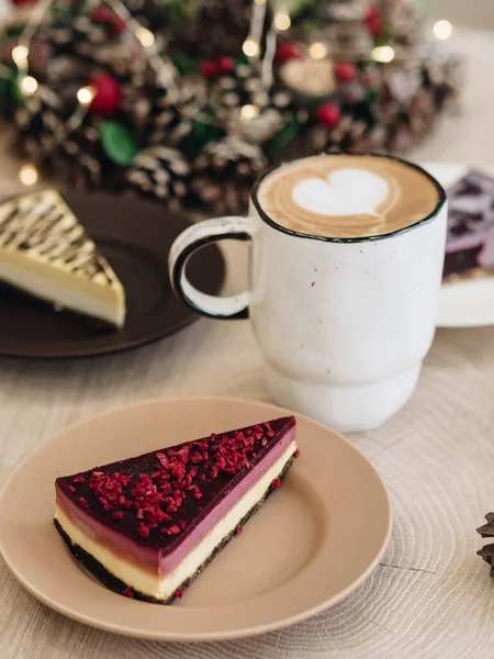 A piece of cake is on a plate. Christmas wreath for home decoration. in the center there is a mug with cappuccino coffee. A heart is drawn on the foam. Warming Christmas drink. Veggie sweets for Christmas.