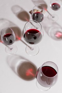 Farklı şekillerde cam kadehler beyaz bir taşın üzerinde duruyor. Kırmızı şarap kutulara dökülür. Arka plana dökülen gözlüklerin uzun gölgeleri. Sommelier. Şarap arıtma. Şarap gelenekleri.