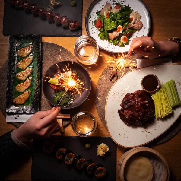 Festive dinner for two. Asian food on the table: Peking duck, rolls, sushi, Chinese dumplings, salad. In the hands of a sparkler. Chinese New Year. There are Chinese chopsticks. Pan-Asian cuisine.