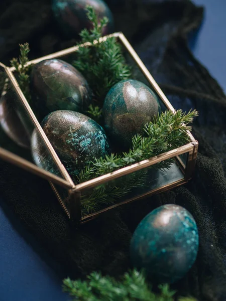 Mother of pearl Easter eggs lie in a glass box and a blue background. There are twigs of juniper between the eggs. An unusual coloring of Easter eggs in the theme of space. Religious holiday Easter.