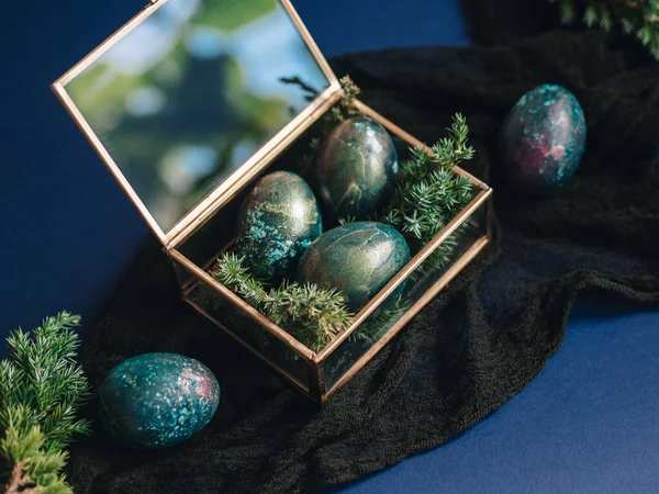 Mother of pearl Easter eggs lie in a glass box and a blue background. There are twigs of juniper between the eggs. An unusual coloring of Easter eggs in the theme of space. Religious holiday Easter.