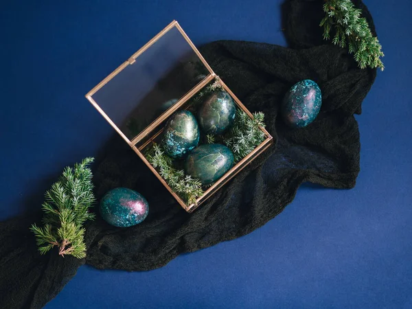 Mother of pearl Easter eggs lie in a glass box and a blue background. There are twigs of juniper between the eggs. An unusual coloring of Easter eggs in the theme of space. Religious holiday Easter.