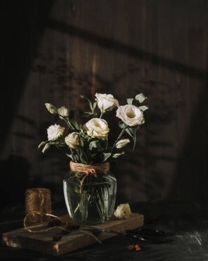 Beyaz çiçekli şeffaf bir vazo güneşte duruyor. Güzel güneş ışığı üzerine düşüyor. Yakınlarda bir makas ve iplik makarası var. Çiçekçi, kesilmiş çiçeklerle ilgileniyor. Bahar havası. Koyu arkaplan.