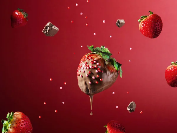 Tangle in chocolate on a red background. Chocolate drips from the berry, pieces of chocolate and strawberries and confectionery sprinkles are falling nearby. Delicious delicacy. Food levitation.
