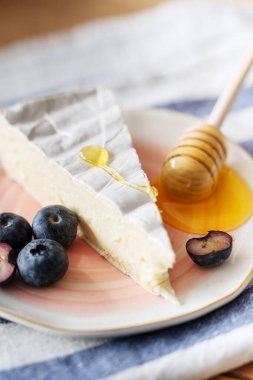 Küçük pembe bir tabakta duran üç köşeli bir peynirli peynir dilimi. Yaban mersiniyle süslenmiş. Yakınlarda bir parça bal var. Peynirli kahvaltı. Şarap çerezi. Camembert peyniri nasıl servis edilir? Genç peynir..